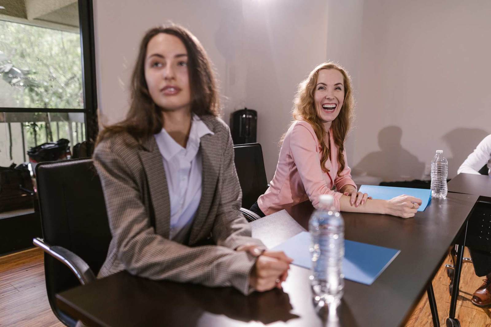 Two women sit at a conference table, one smiling broadly, with water bottles and folders in front of them