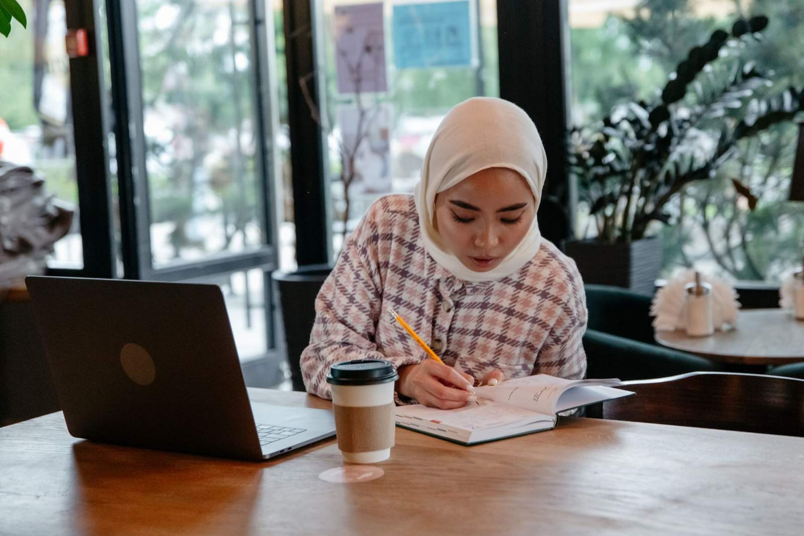 A woman in a hijab writes in a notebook beside an open laptop and a coffee cup at a café table