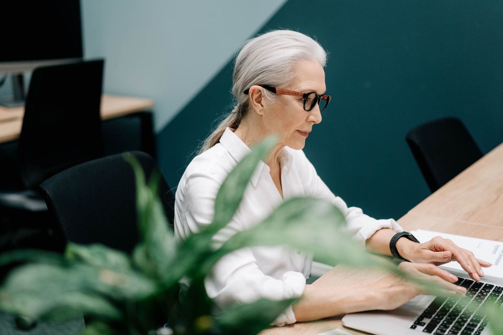 A woman with gray hair and glasses works at a laptop at a conference table in an office, a plant in the foreground