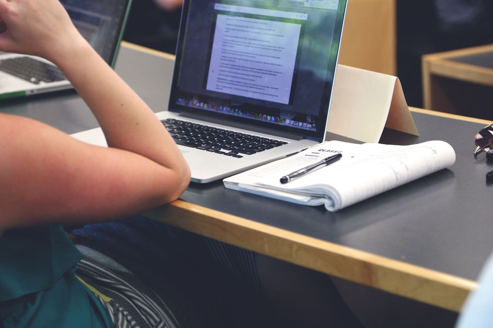 A person works at a laptop displaying a document, with an open notebook, pen, and sunglasses on the desk beside them, seen from over their shoulder