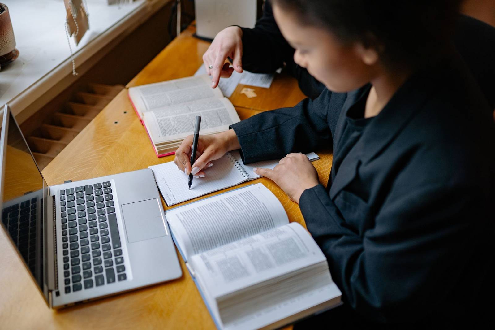 Two people sit at a desk poring over open reference books and handwritten notes beside a laptop, one pointing to a passage on the page