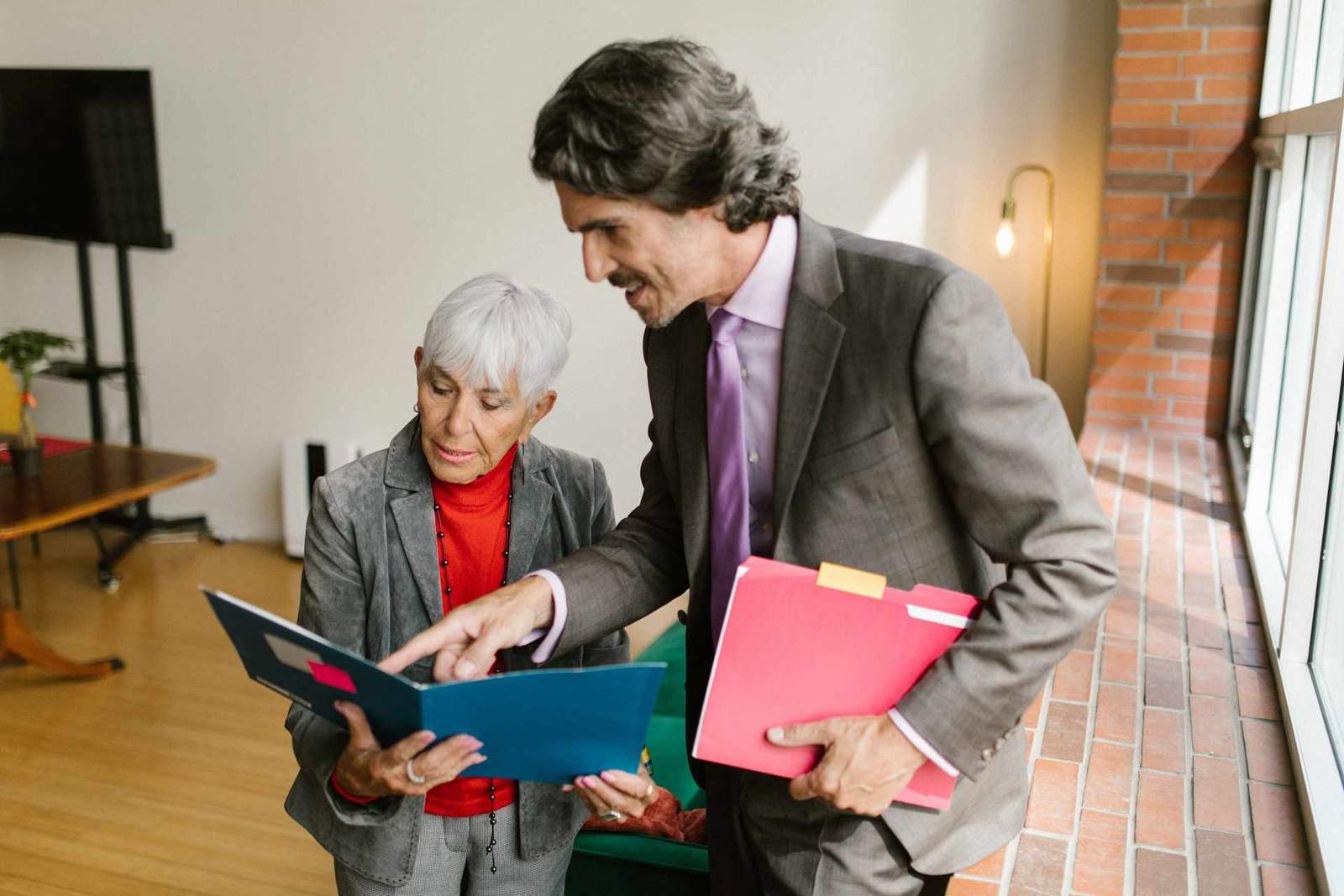 An older woman and a man in business suits stand reviewing an open folder together, one pointing at a page
