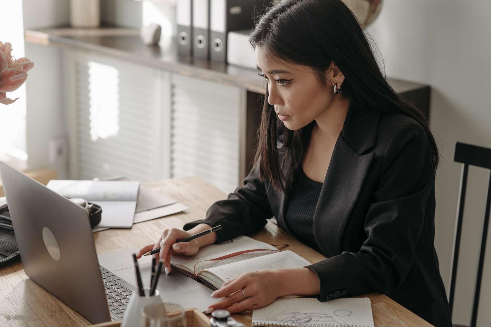 A woman in a blazer writes in a notebook while reviewing a laptop screen at an office desk