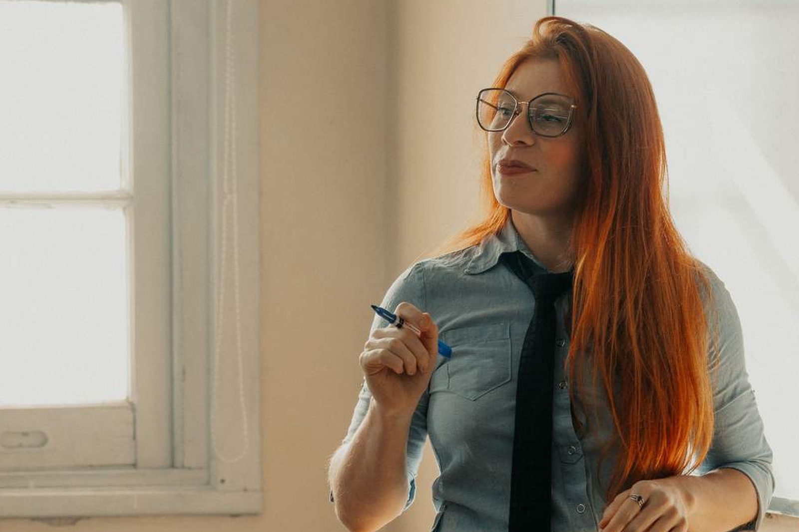 A woman wearing glasses holds a pen and looks thoughtfully to the side near a sunlit window