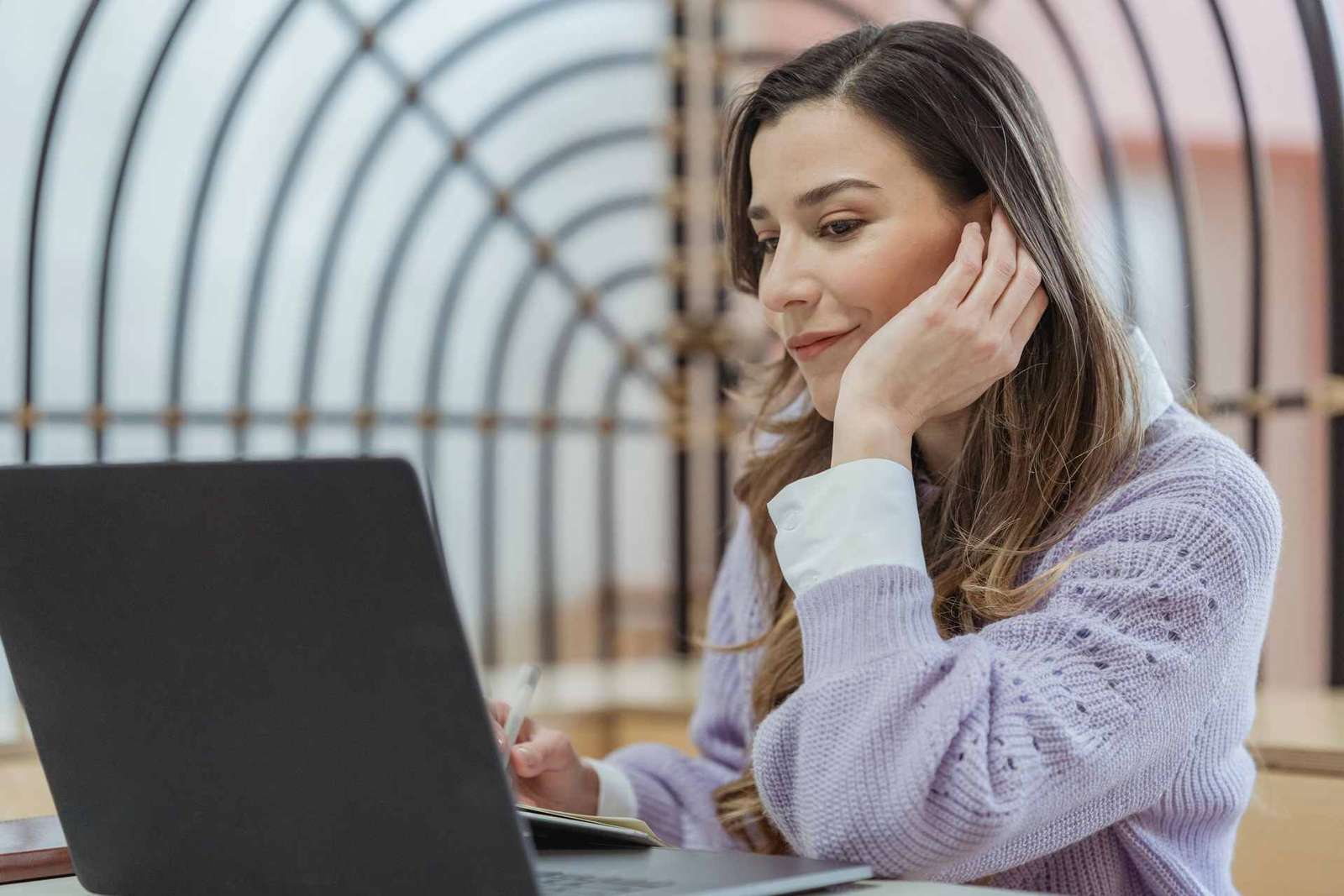 A woman rests her chin in her hand while reading something on a laptop beside an open notebook