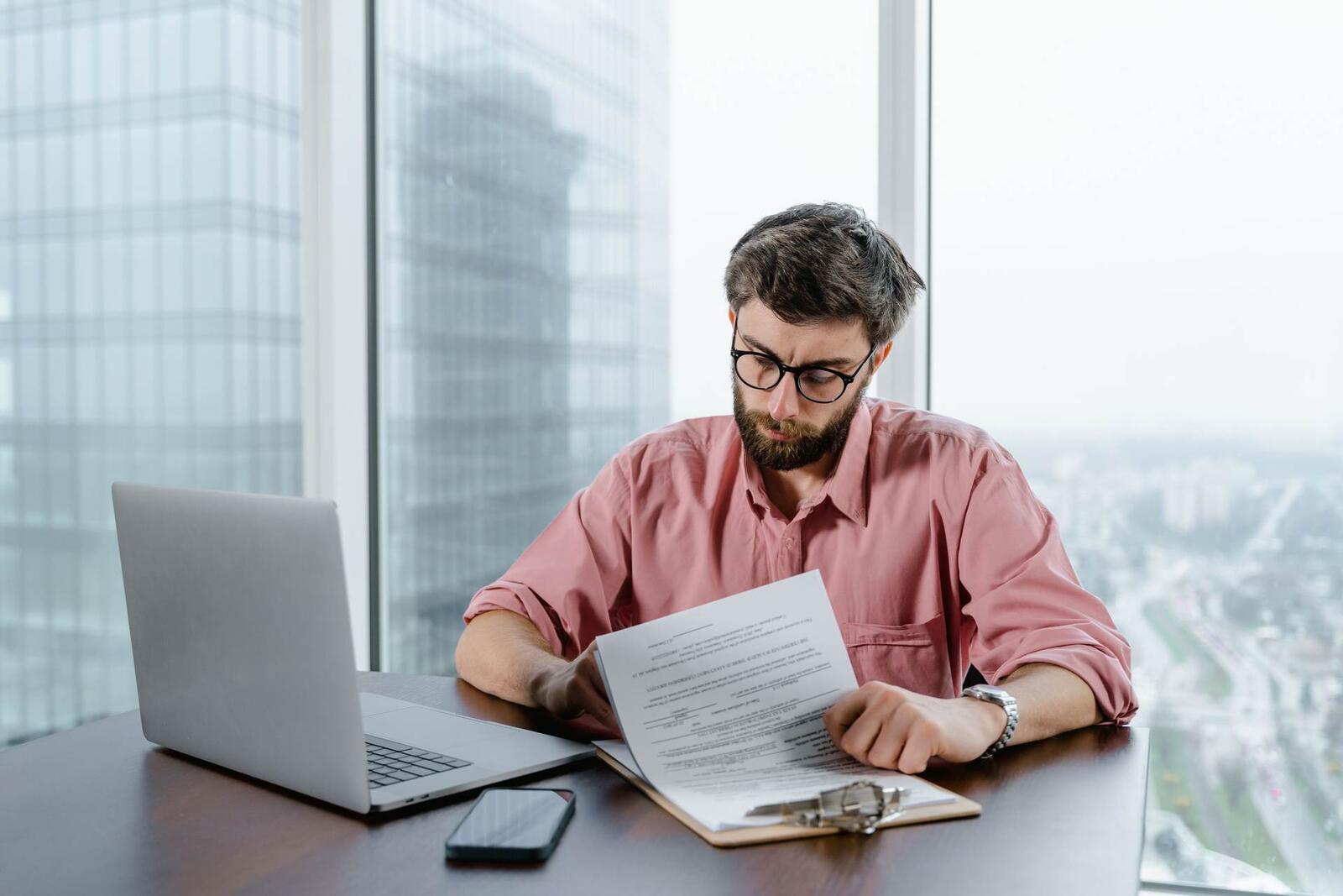 A bearded man in glasses and a pink shirt reviews a clipped document beside a laptop in a high-rise office