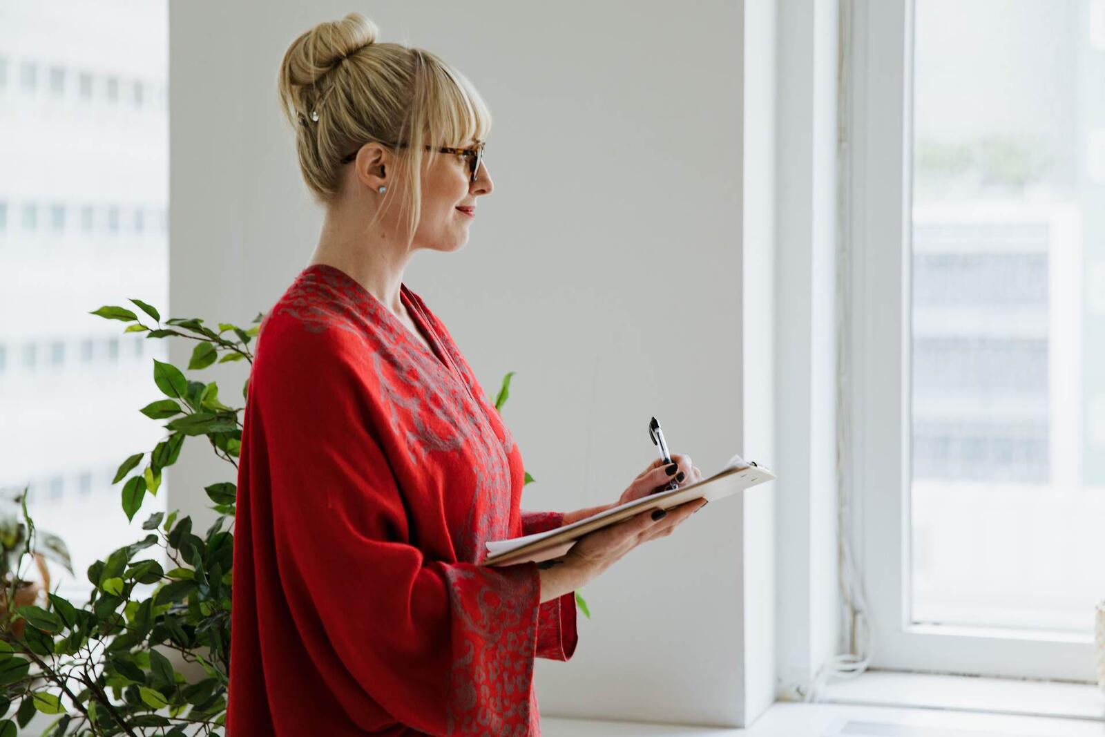A woman in a red patterned wrap writes on a clipboard while looking out a tall office window, blurred city office buildings visible beyond the glass