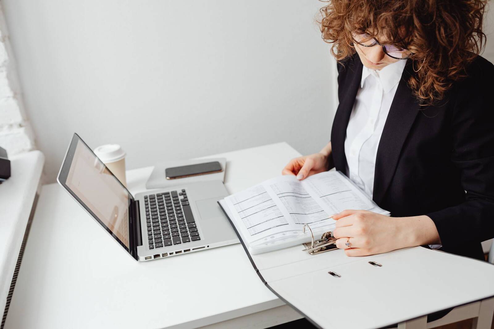 A woman in a black blazer reviews a binder of tabulated data beside an open laptop at a white desk