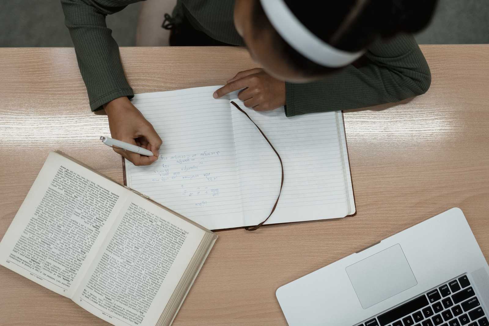 An overhead view of a person wearing headphones cross-referencing an open dictionary while writing in a notebook beside a laptop
