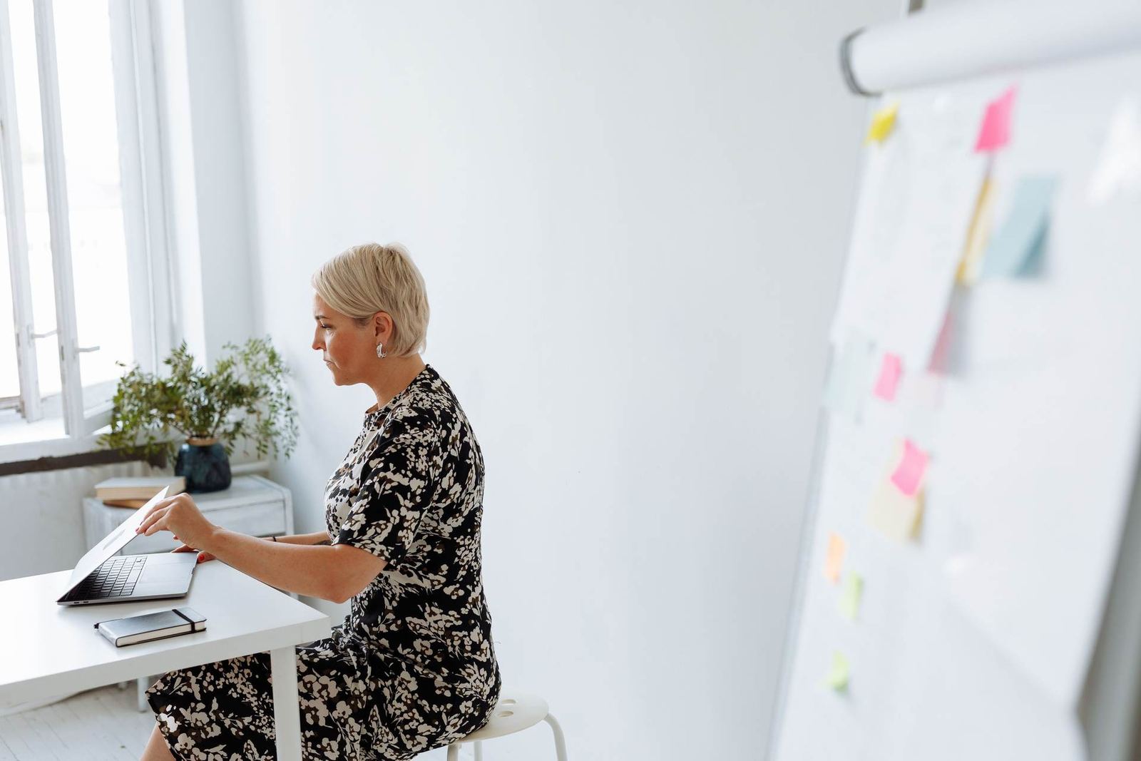 A woman in a floral dress sits at a desk working on a laptop beside a window, with a whiteboard covered in colorful sticky notes in the background