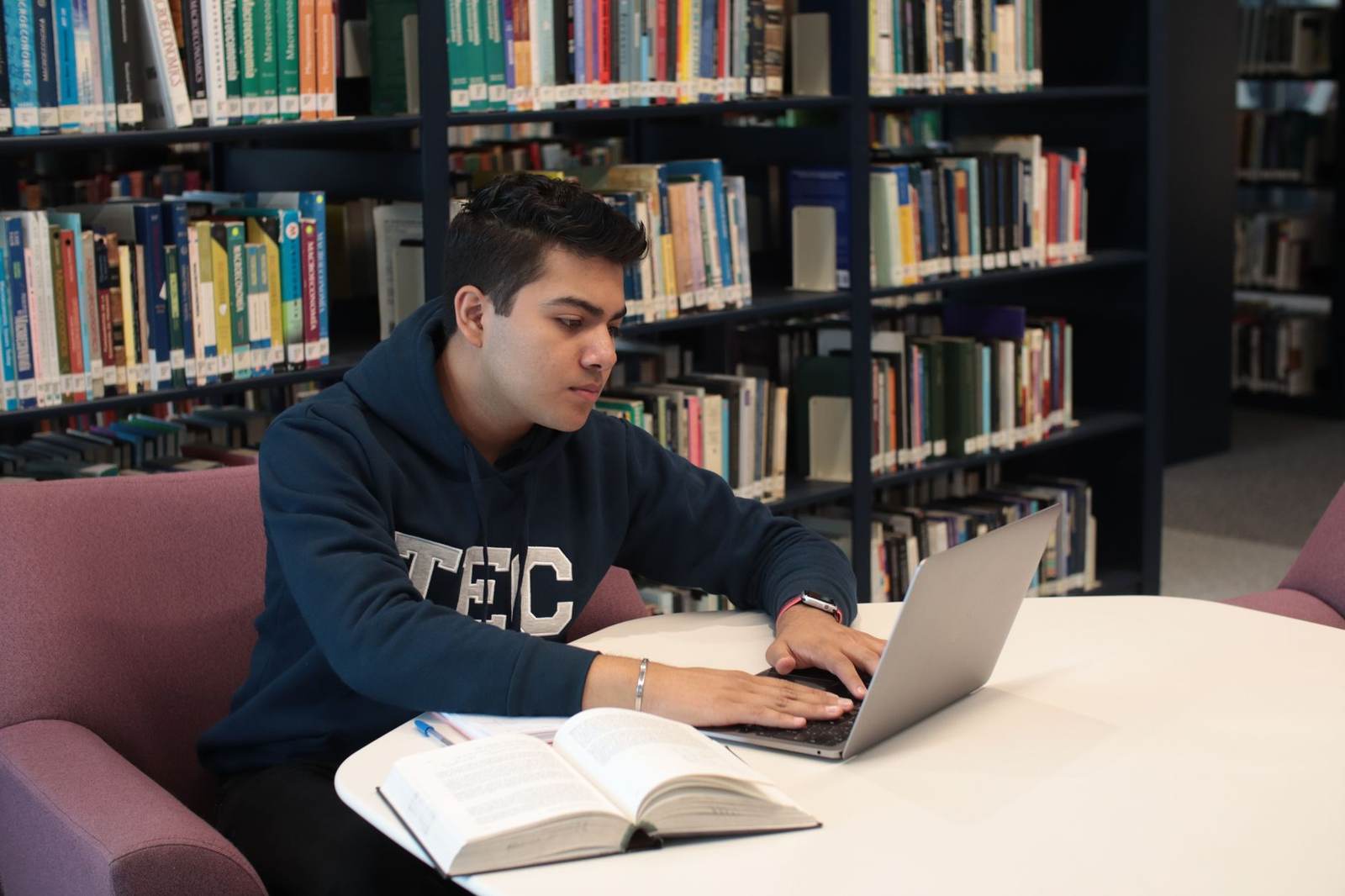 A student works at a laptop beside an open textbook at a library table, rows of bookshelves in the background