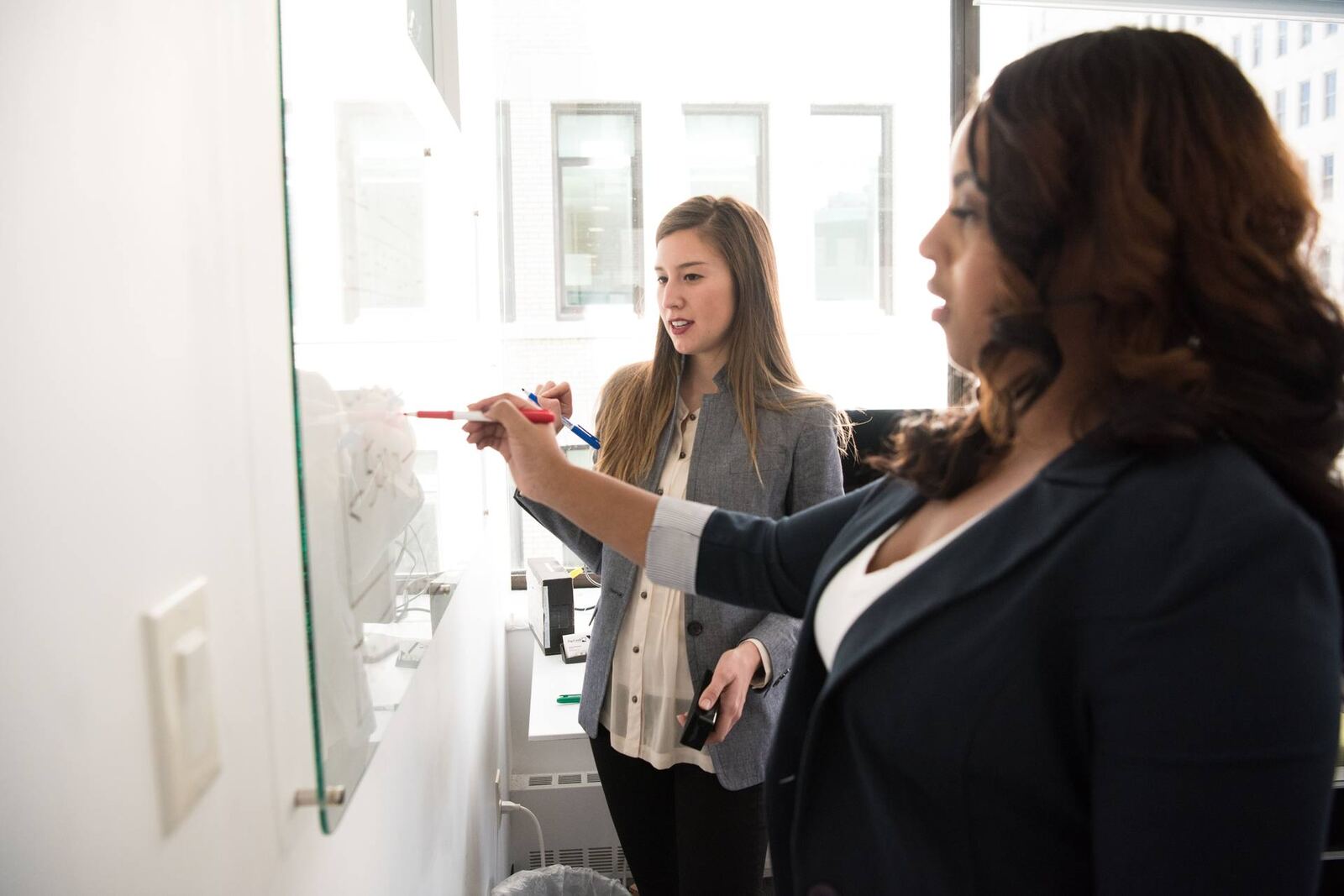 Two colleagues in blazers confer at a glass whiteboard in a bright office, one pointing with a marker toward faint notes