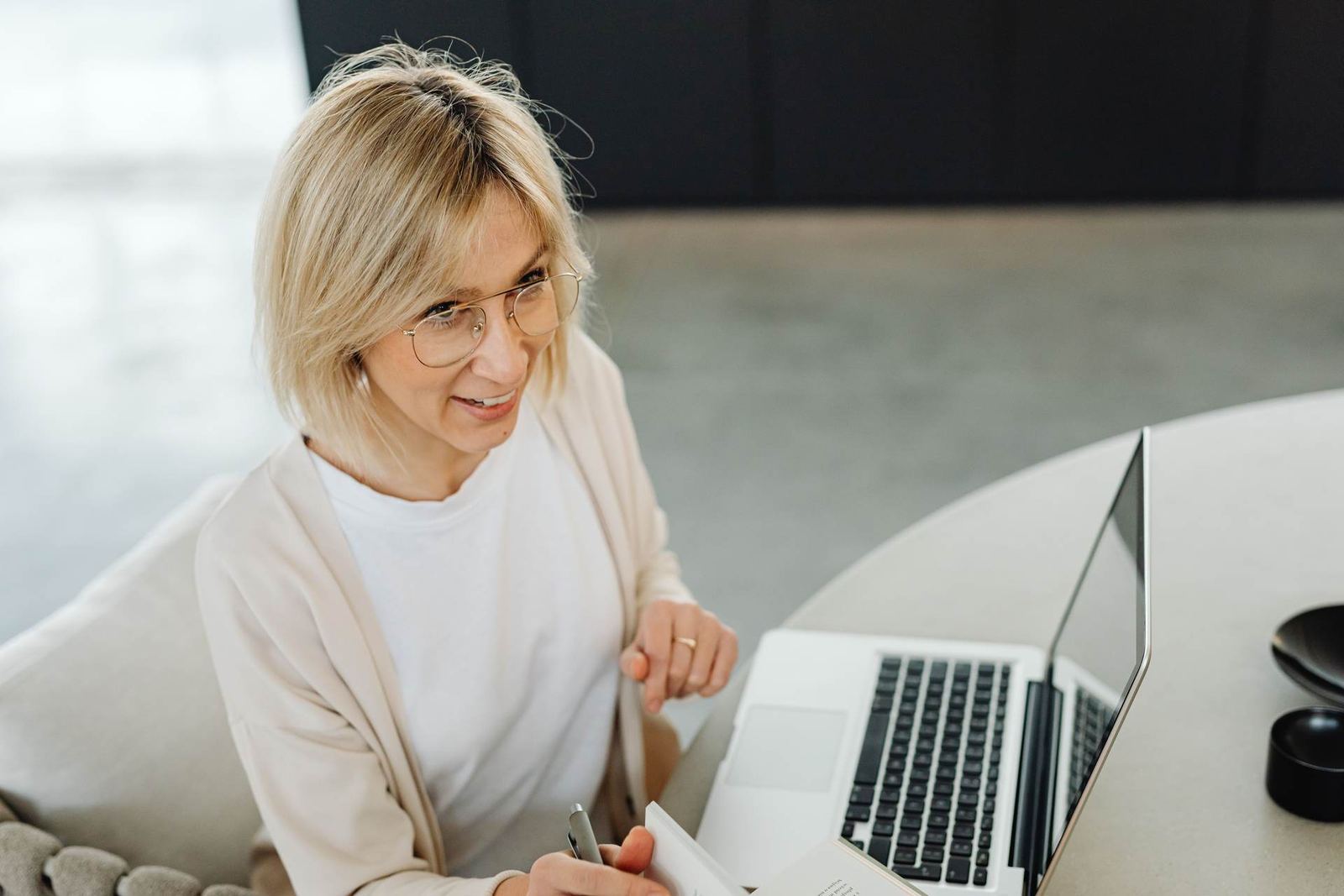 A woman wearing glasses smiles while writing in a notebook beside an open laptop