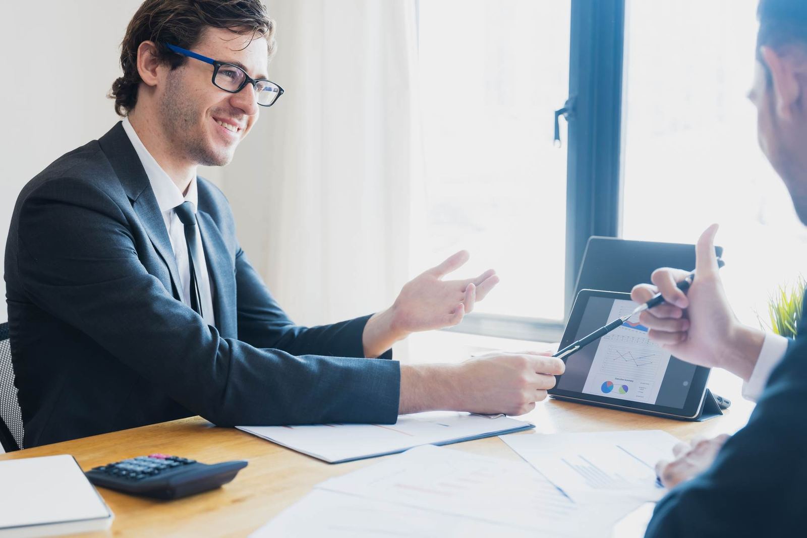 Two men in suits sit at a desk, one gesturing while the other points a pen at a tablet showing a chart