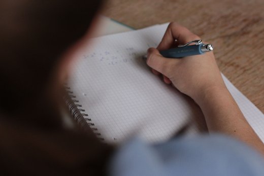 A close-up of a hand annotating handwritten notes in a spiral notebook with a pen, seen over the writer&rsquo;s shoulder