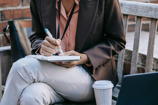 A person writes in a notebook resting on their lap, a lanyard visible at the collar, a coffee cup and laptop nearby