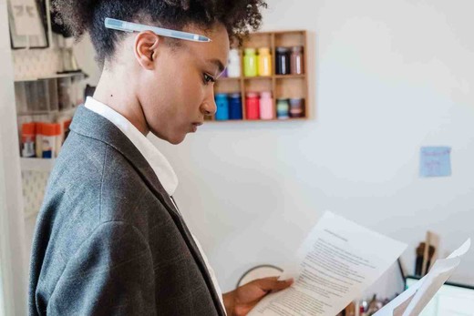 A woman in a blazer reads a printed document closely while standing in a home office, a pen tucked behind her ear