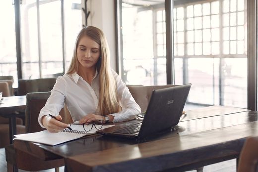 A woman in a white shirt writes in a notebook at a wooden table beside an open laptop