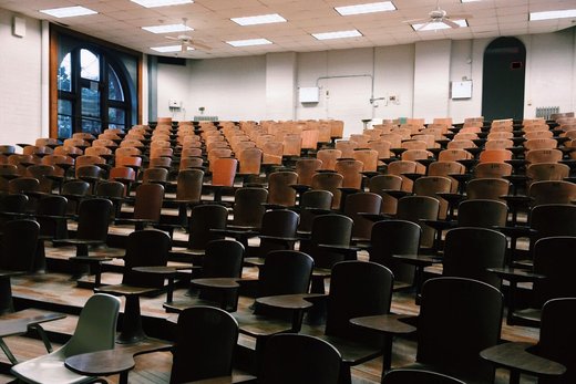 Rows of empty vintage lecture hall seats with tablet armrests, ceiling fans, and arched windows in the background
