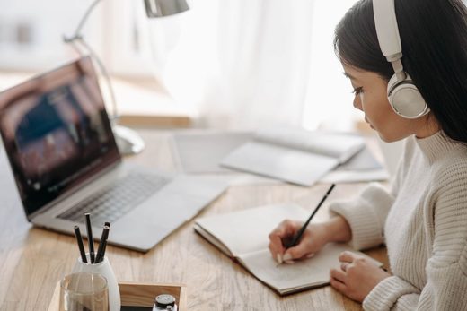 A woman wearing headphones writes in a notebook beside an open laptop at a desk