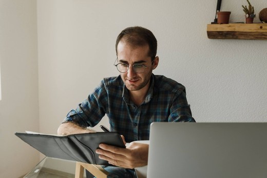 A man at a home desk reviews a document in an open folder while writing, with a laptop open in front of him