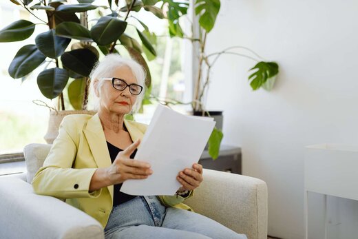An older woman in a yellow blazer and glasses reads a stack of papers in an armchair surrounded by houseplants