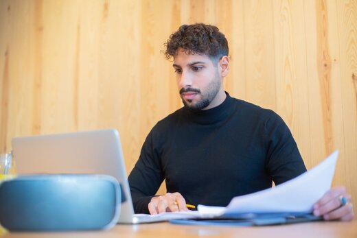A man in a black turtleneck reviews papers at a laptop against a wood-paneled wall