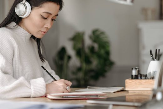 A woman wearing headphones writes in a notebook at a desk beside an open laptop and notepad