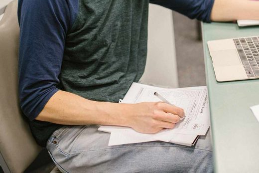 A hand writes on a printed worksheet beside an open laptop on a desk
