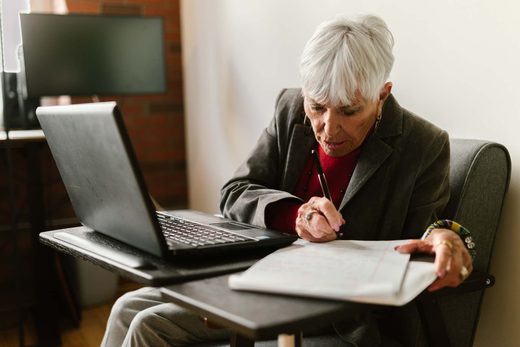 An older woman in a blazer reviews handwritten notes beside an open laptop