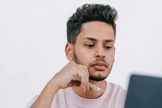A man rests a pen against his chin, reading closely from a laptop screen