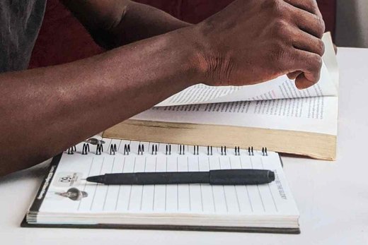 A close-up of a person's hand turning a page in a hardcover book beside a spiral notepad and pen