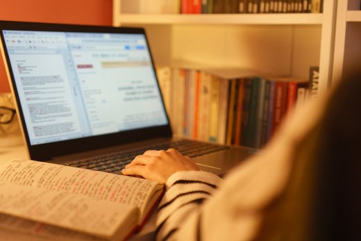 A person types on a laptop beside an annotated notebook, in front of a bookshelf, seen over their shoulder