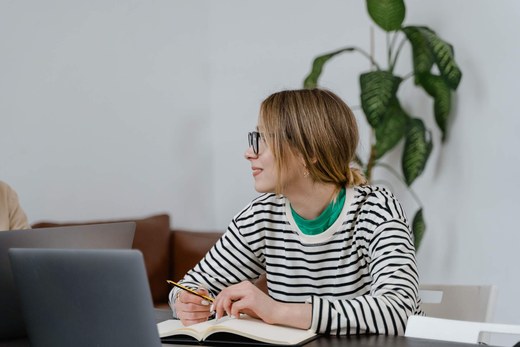 A woman with a notebook and pencil sits at a table with open laptops during a discussion