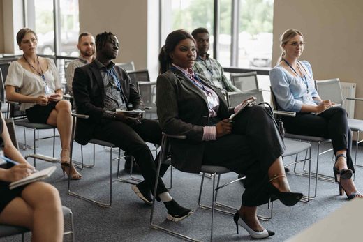 A row of seminar attendees wearing lanyards sit taking notes while listening in a bright meeting room