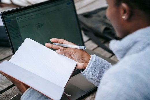A person reviews a handwritten notebook with a pen in hand beside an open laptop showing an email inbox