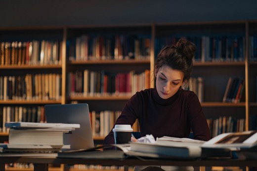 A woman studies late at night at a library table stacked with books and a laptop, rows of bookshelves behind her