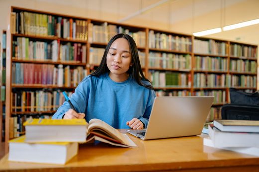 A student writes in a notebook at a library table, an open laptop and stacked reference books in front of her, bookshelves in the background