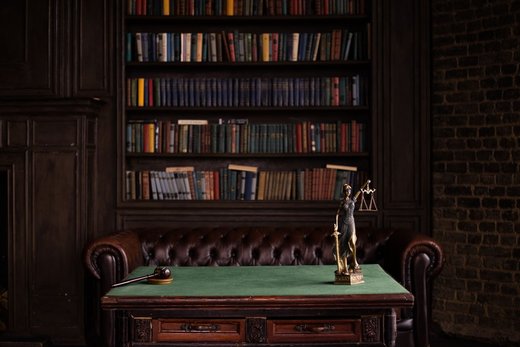 A gavel and a small blind-justice statuette holding scales, on a green desk pad in front of a dark wood bookshelf lined with law books