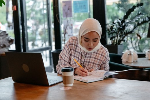 A woman in a hijab writes in a notebook beside an open laptop and a coffee cup at a café table
