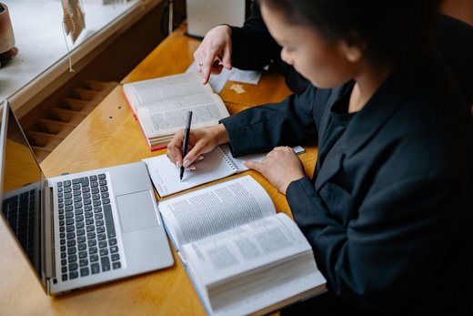 Two people sit at a desk poring over open reference books and handwritten notes beside a laptop, one pointing to a passage on the page