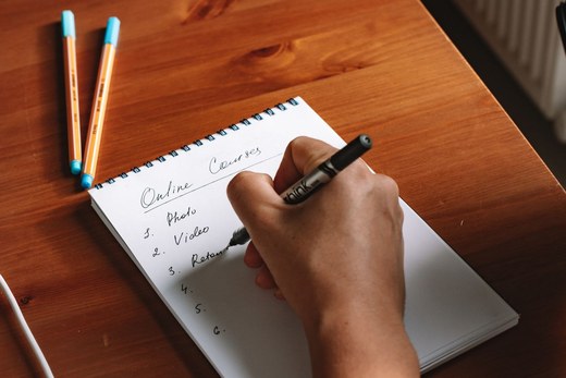 A hand writes a numbered list on a spiral notepad with a marker, two pens resting nearby on a sunlit wooden desk