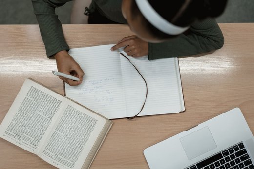 An overhead view of a person wearing headphones cross-referencing an open dictionary while writing in a notebook beside a laptop