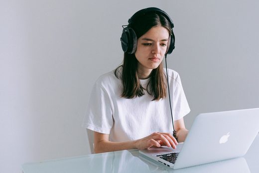 A person wearing headphones sits at a desk typing on a laptop against a plain wall