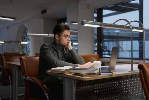 A person at a desk in a dim office reads from a stack of open books while typing on a laptop, a coffee cup nearby