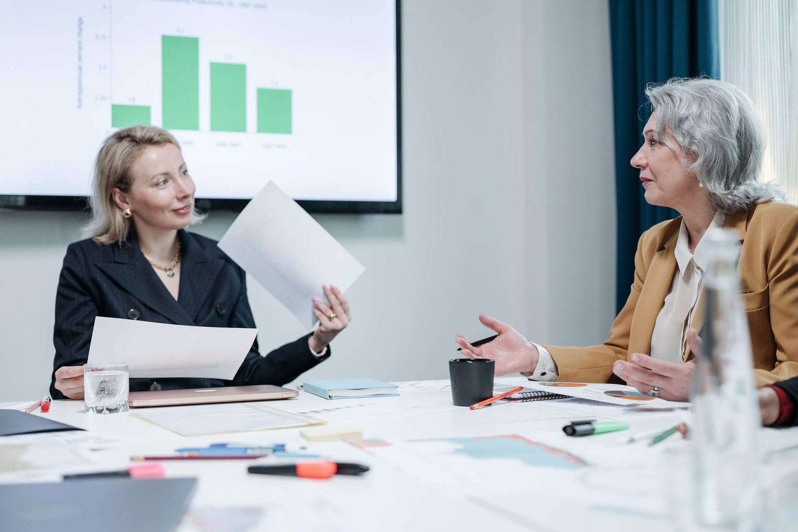 Two colleagues talk over papers at a table with a bar chart shown on a screen behind them