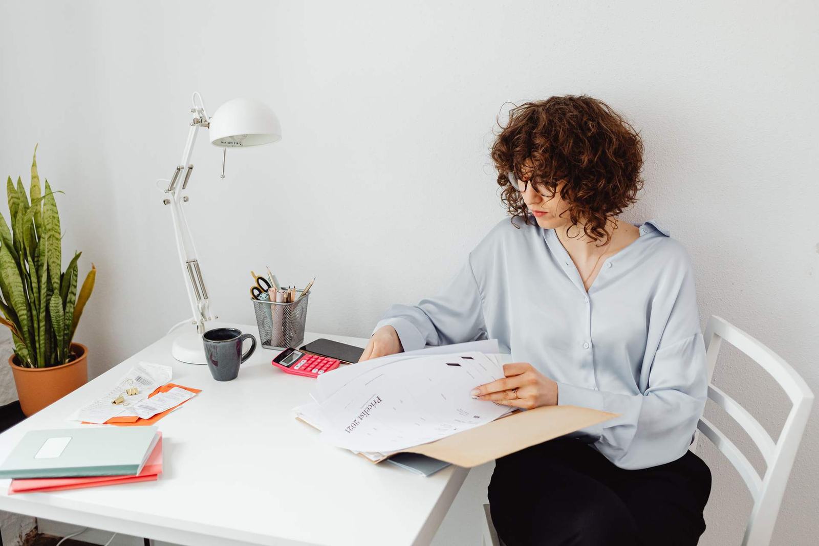 A woman in a light blue blouse reviews printed documents and a folder at a bright desk with a lamp and a calculator