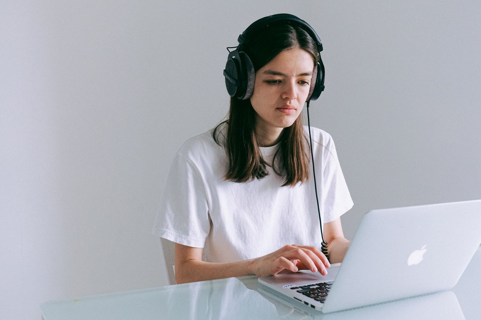 A person wearing headphones sits at a desk typing on a laptop against a plain wall