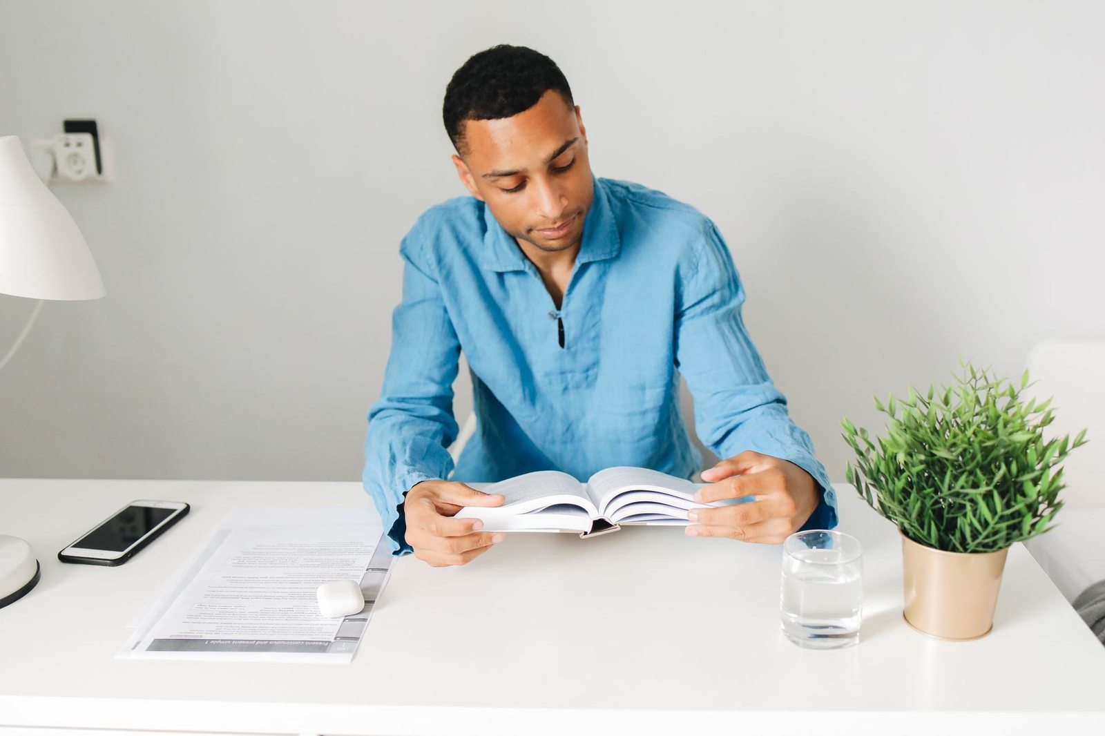 A man in a blue shirt sits at a white desk reading closely from an open book, a phone and potted plant nearby