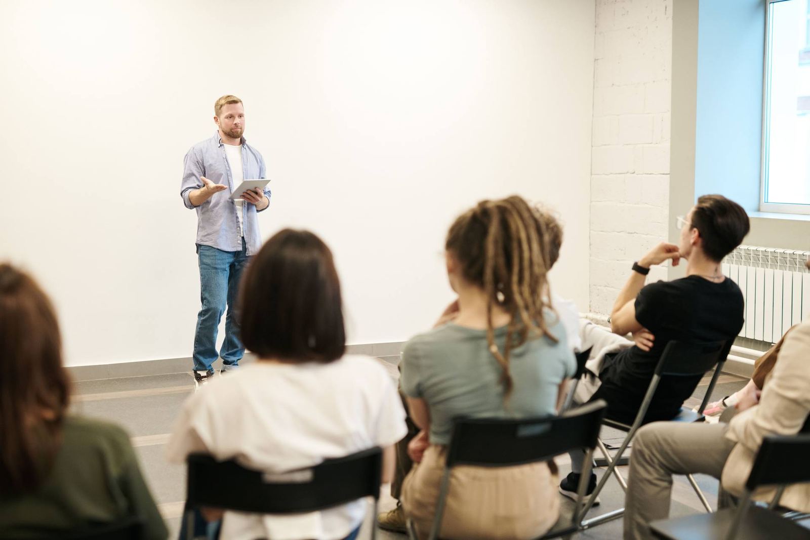 A man holds a tablet and gestures while addressing a small seated group in a meeting room